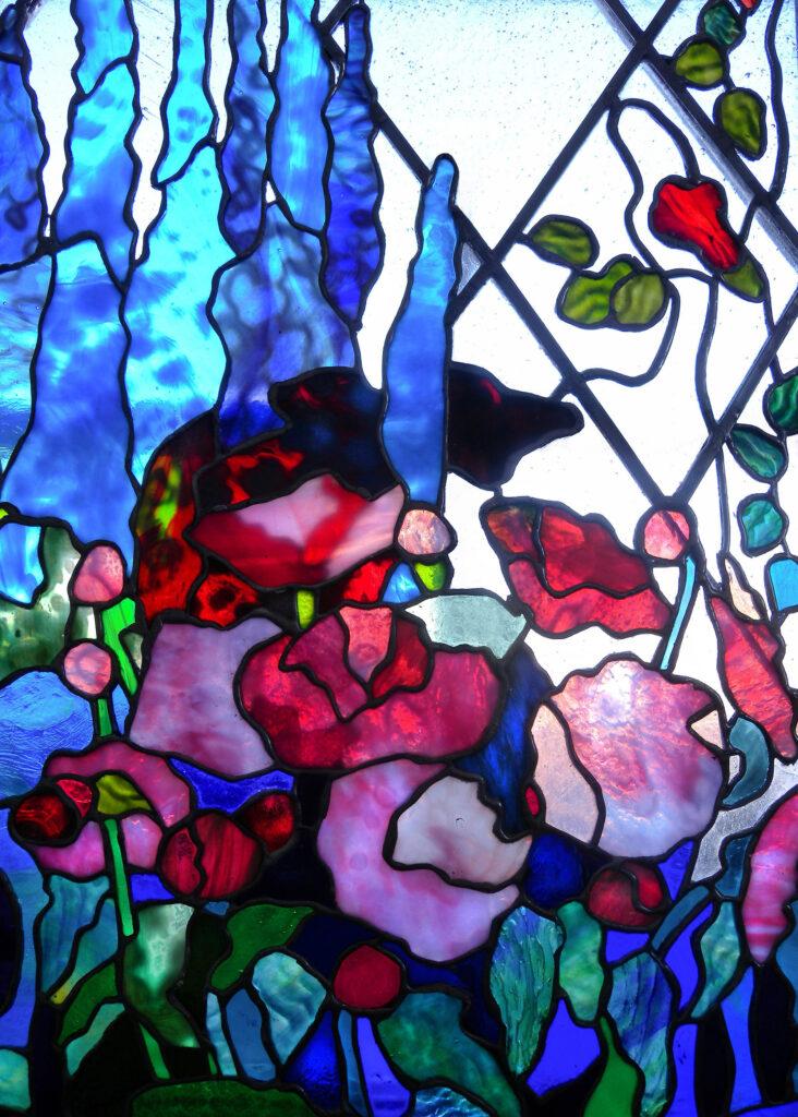 Stained glass window, Tiffany style, featuring red poppies, blue foxglove flowers, and green leaves. Against frosted, diamond-patterned glass background. It is a custom garden scene made by Alexandra Puzyn.