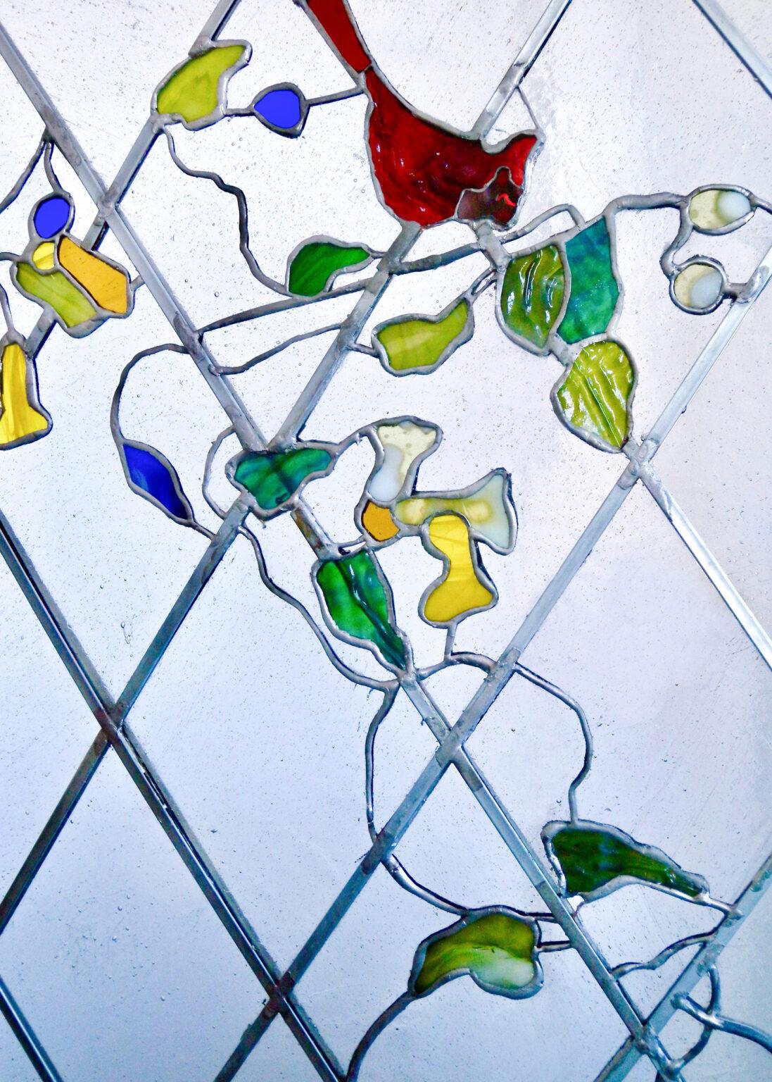 Stained glass leaded panel featuring a decorative floral design with flowing shapes of vines, leaves, and yellow flowers with a red bird. The organic lines of the vines and stylized flowers are set against a background of clear, textured glass in a diamond pattern. Made and designed by stained glass artist Aleksandra Puzyn.