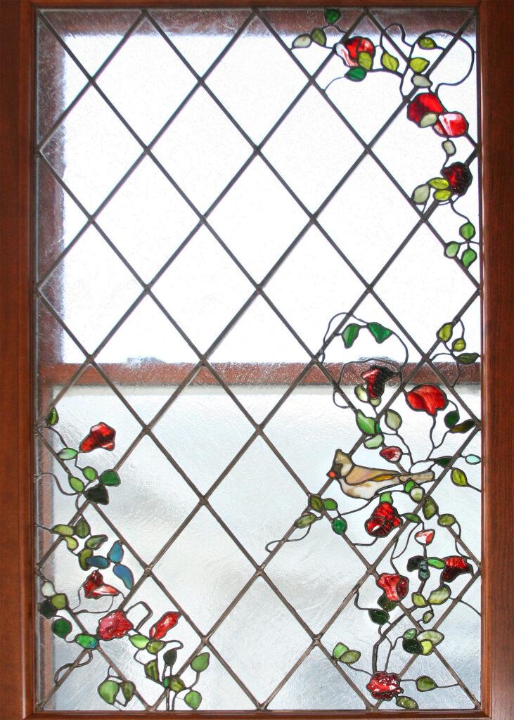 Custom-made leaded stained glass window panel featuring a naturalistic design of a Cardinal bird, and a winding vine of red roses. The background uses a diamond pattern of clear textured glass. It is an intricate Tiffany-style floral design made and fabricated by Alexandra Puzyn, displayed in a residential window.