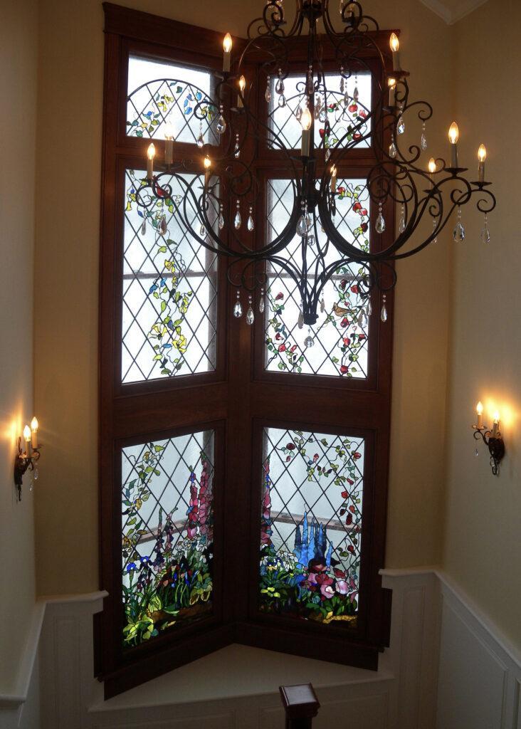Custom-made stained glass window installation in a residential house. It is a two-story high window that features a diamond-pattern leaded design with colorful botanical motifs, including vines, roses, and birds. The lower panels depict elaborate garden scenes with various types of flowers. The window is set in rich, dark wood. It is Alexandra Puzyn artwork.