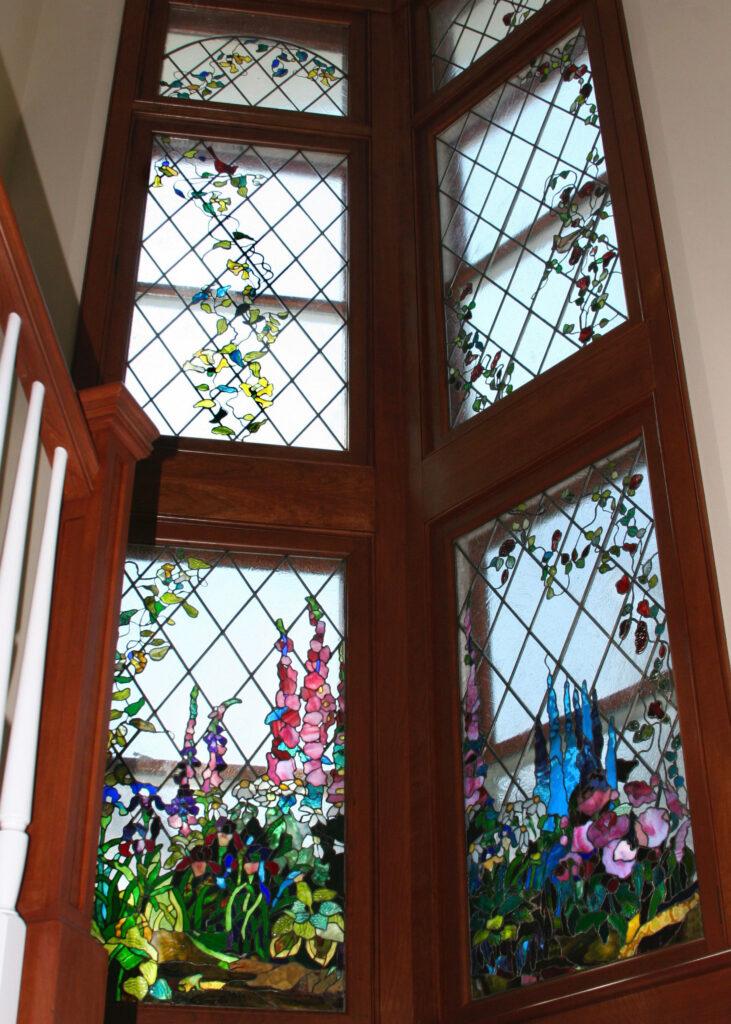 Custom-made stained glass in a residential house. A two-story high window features a colorful botanical motifs, including vines, roses, and birds with diamond-pattern leaded design. The lower panels depict elaborate garden scenes with various types of flowers. The window is set in rich, dark wood. It is Alexandra Puzyn artwork.
