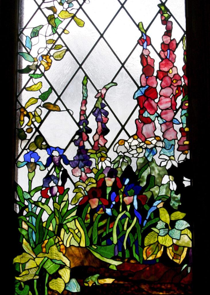 Stained glass window featuring an intricate design of a garden with vivid and colorful flowers. Created in the Tiffany style, this piece uses opalescent and favrile glass with varied textures. The technique involves using Cooper foil and lead came, made by artist Alexandra Puzyn.