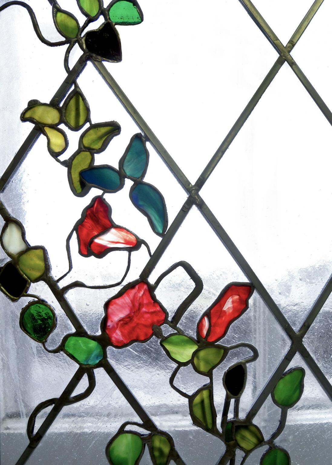 Leaded stained glass window panel featuring a floral design in the Art Nouveau style. The roses are rendered in vibrant reds, with green and blue-green leaves and twisting vines set against a background of clear, textured glass in a diamond pattern. The organic lines of the vines and stylized flowers are made and designed by stained glass artist Aleksandra Puzyn.