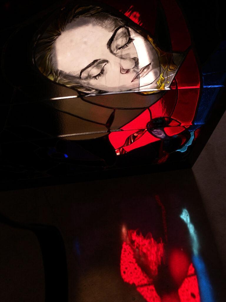Stained glass portrait of a woman's face, shown with her eyes closed, amidst vibrant red and blue glass. Intense red hues create a dramatic interplay of light and shadow, and cast vibrant reflections onto the surface below.
